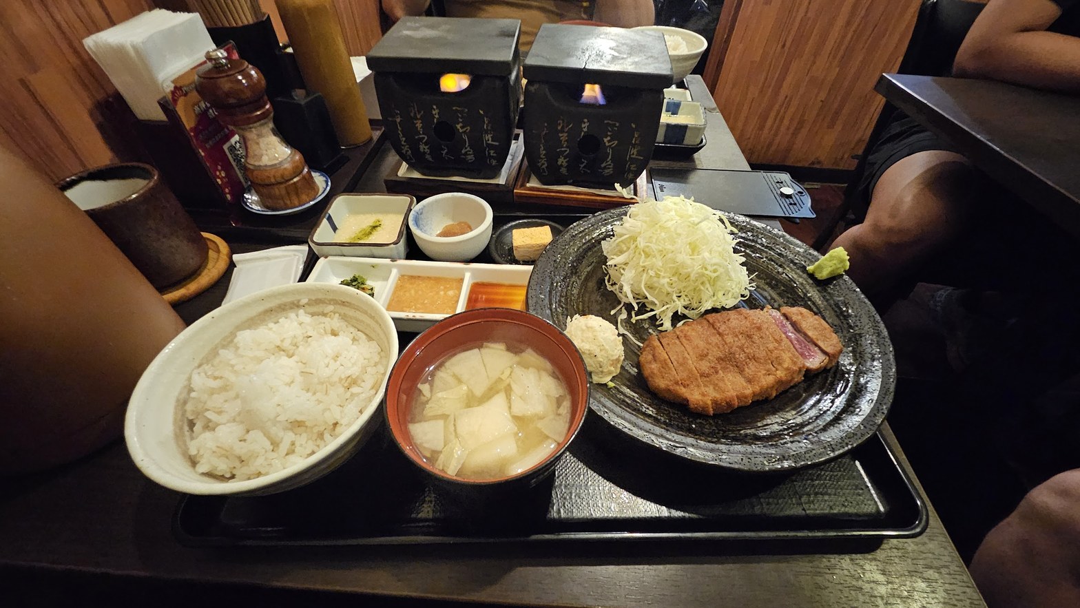 黒いトレーに乗った牛かつ定食と2台の石焼きプレート、炎が灯り奥席に複数のセットが並ぶ