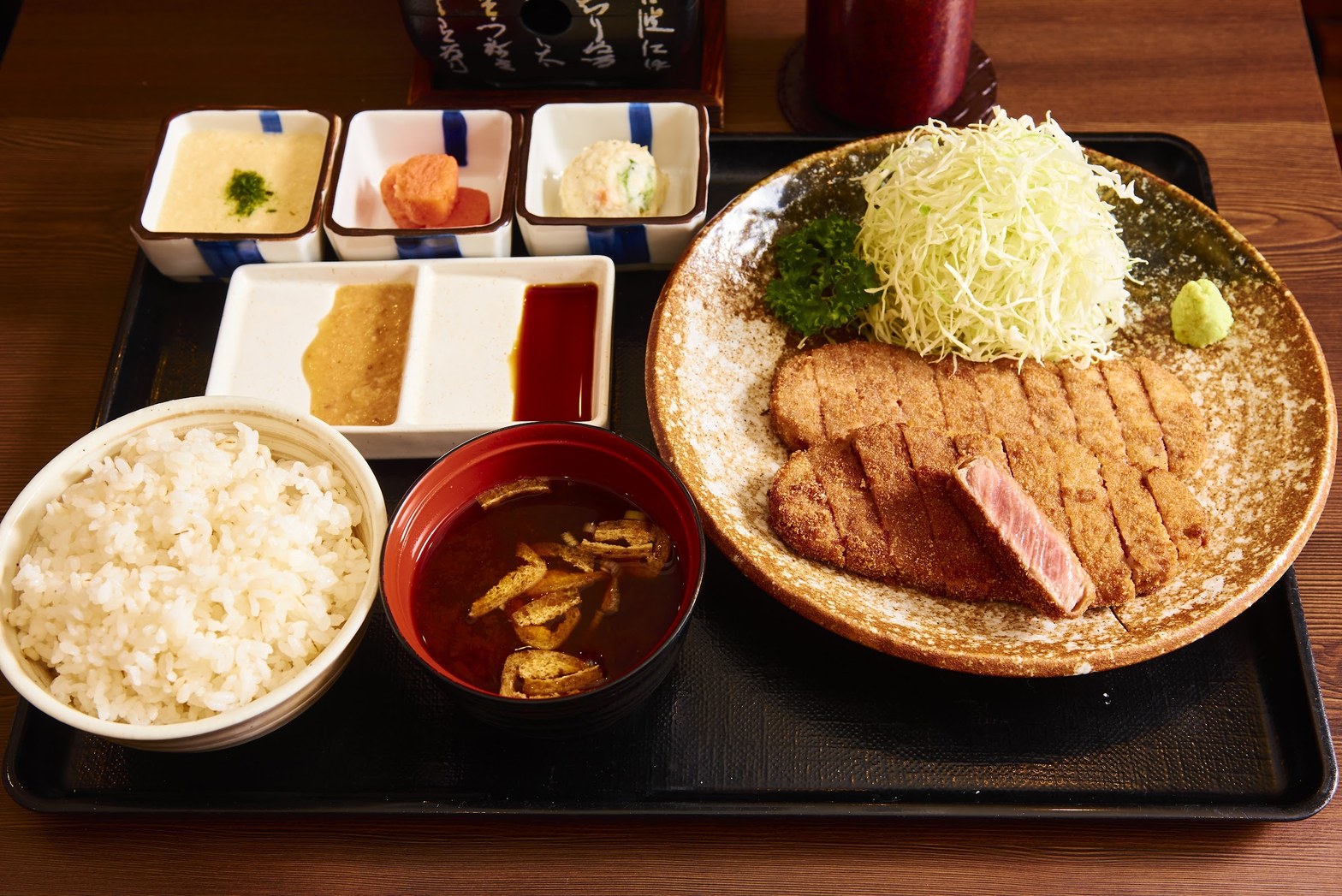 黒いトレーに乗った牛かつ定食——石皿の牛かつ・麦飯・赤い椀の味噌汁・小鉢3種・2種のタレが並ぶ